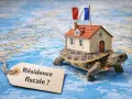 Une tortue porte une maison avec un drapeau français, sur une carte du monde. Une étiquette demande : "Résidence fiscale ?".
