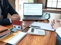 Réunion pour discuter de la faute du mandataire de gestion locative : maison modèle, calculatrice, documents et ordinateur portable sur un bureau en bois.