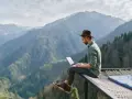 Un homme en chapeau travaille sur son ordinateur portable, profitant du télétravail à l'étranger avec une vue imprenable sur les montagnes.