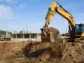 Pelleteuse jaune en action, modifiant le niveau du terrain naturel sur un chantier. Une chargeuse et une grue sont visibles en arrière-plan.