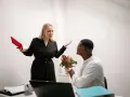 Une femme en colère, tenant un livre rouge, semble se faire insulter au travail par un collègue qui a les mains jointes.