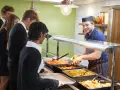 Des élèves font la queue pour le déjeuner à la cantine. Un employé souriant sert des légumes. Un contrat extra restauration est en cours de discussion.