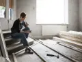 Femme en salopette assise sur des marches, tablette à la main, réfléchissant aux travaux de second œuvre. Matériaux de construction et outils au sol.