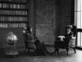 Deux personnes assises dans des fauteuils en cuir, une femme en robe longue et un homme en costume, discutent dans une bibliothèque. Scène évoquant des témoignages procédure en départage prud'hommes.
