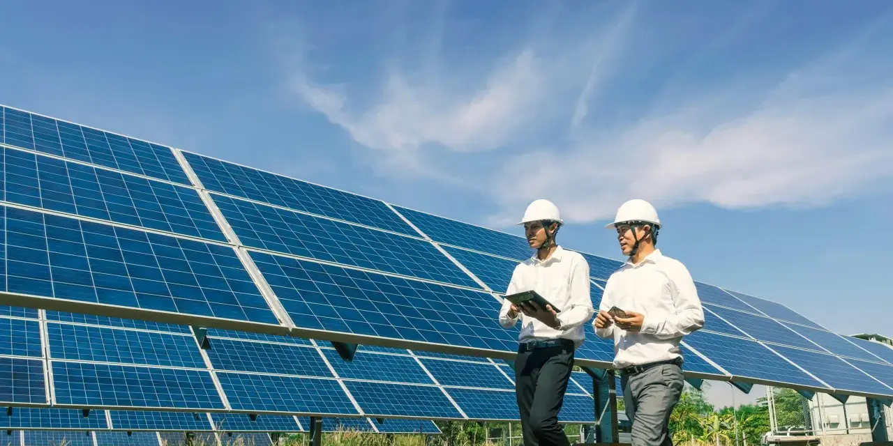 Deux hommes en casque blanc inspectent un champ de panneaux solaires, discutant d'un bail emphytéotique photovoltaïque sous un ciel bleu.