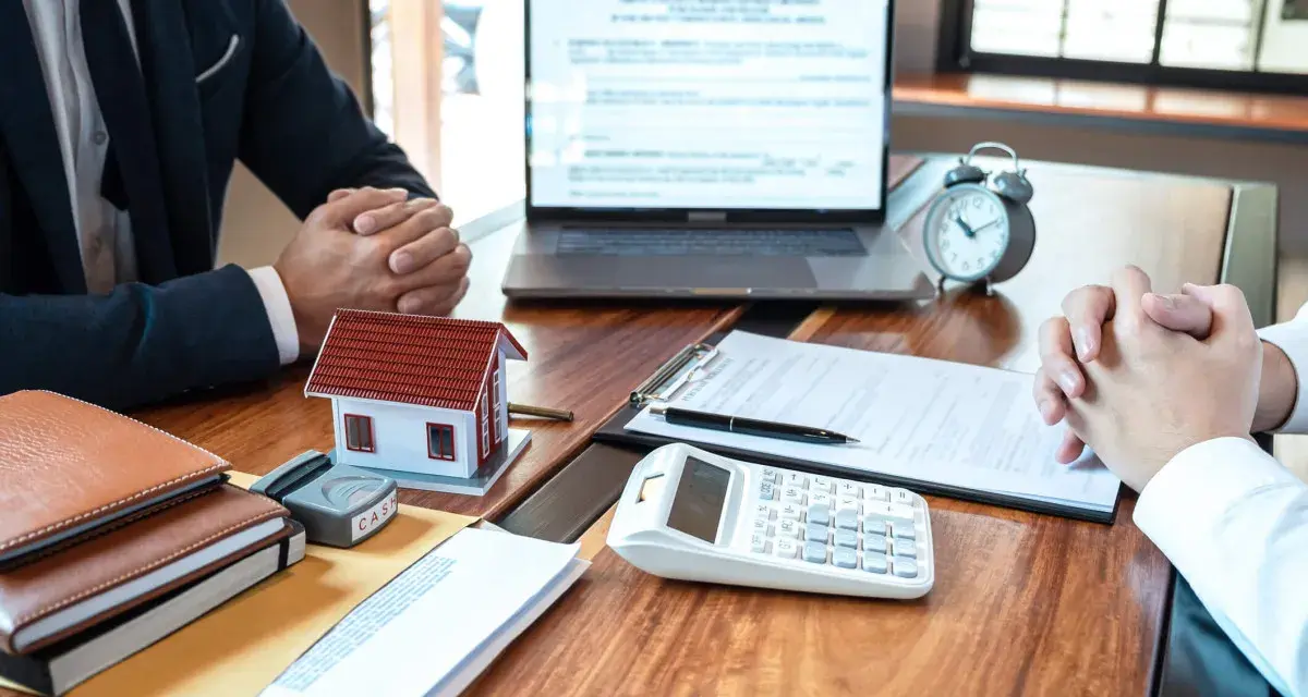 Réunion pour discuter de la faute du mandataire de gestion locative : maison modèle, calculatrice, documents et ordinateur portable sur un bureau en bois.