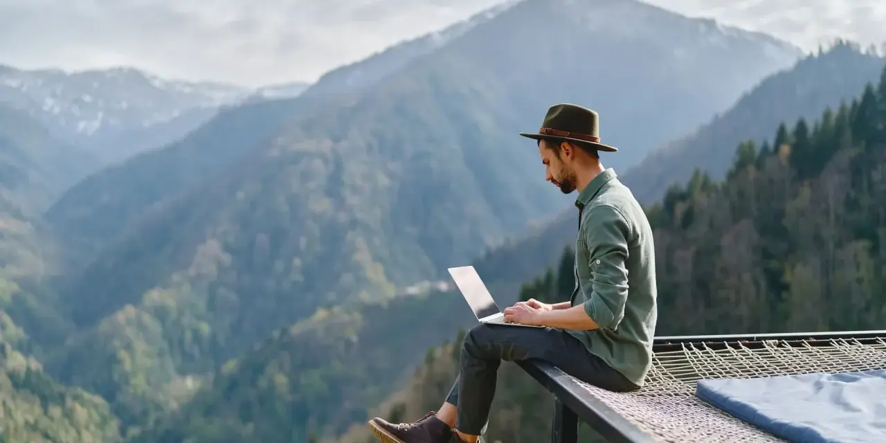 Un homme en chapeau travaille sur son ordinateur portable, profitant du télétravail à l'étranger avec une vue imprenable sur les montagnes.