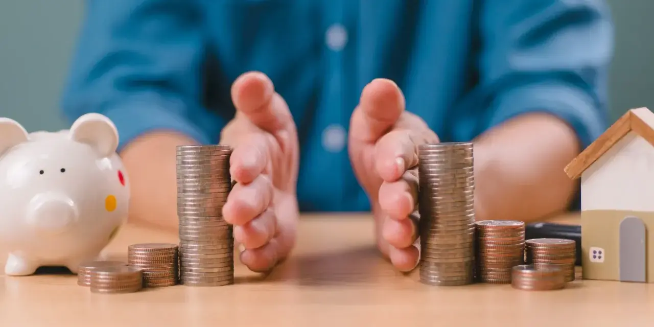 Mains protégeant des piles de pièces, symbolisant la gestion financière et la **desolidarisation pret immo**. Une tirelire et une maison miniature sont visibles.