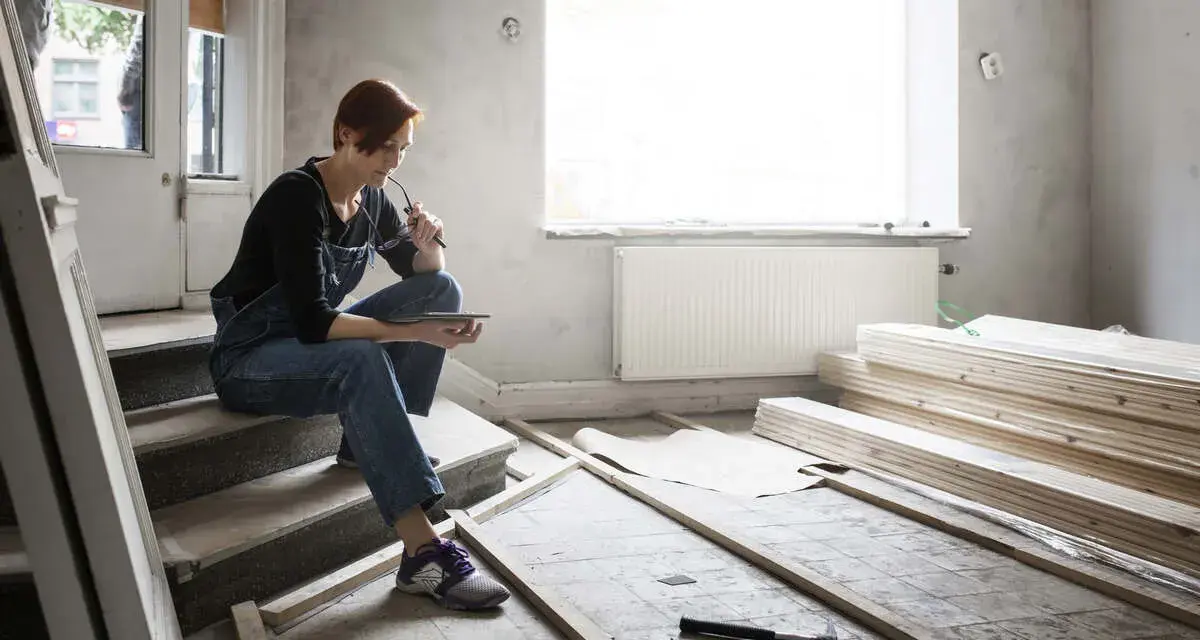 Femme en salopette assise sur des marches, tablette à la main, réfléchissant aux travaux de second œuvre. Matériaux de construction et outils au sol.
