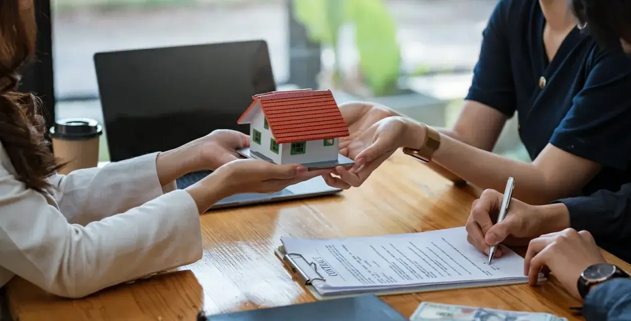 Une maison miniature est remise, un contrat est signé, et de l'argent est posé sur la table, suggérant une transaction immobilière et un chèque de caution.