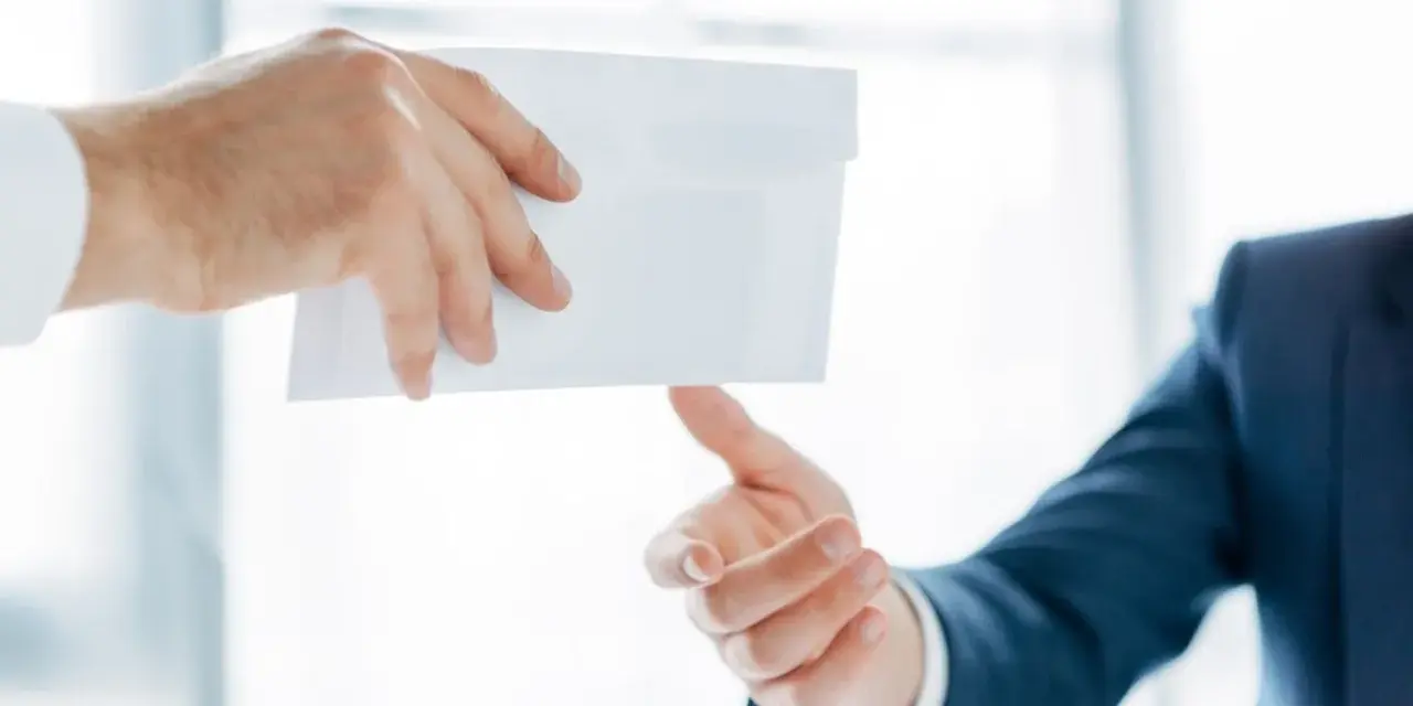Main d'un homme en costume remettant une enveloppe blanche à une autre main. Peut-être une mise en demeure URSSAF avec un délai de paiement.