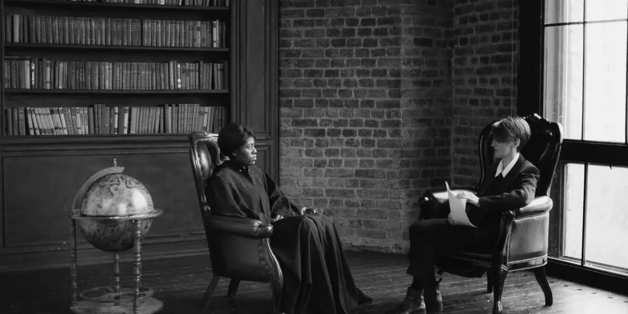 Deux personnes assises dans des fauteuils en cuir, une femme en robe longue et un homme en costume, discutent dans une bibliothèque. Scène évoquant des témoignages procédure en départage prud'hommes.