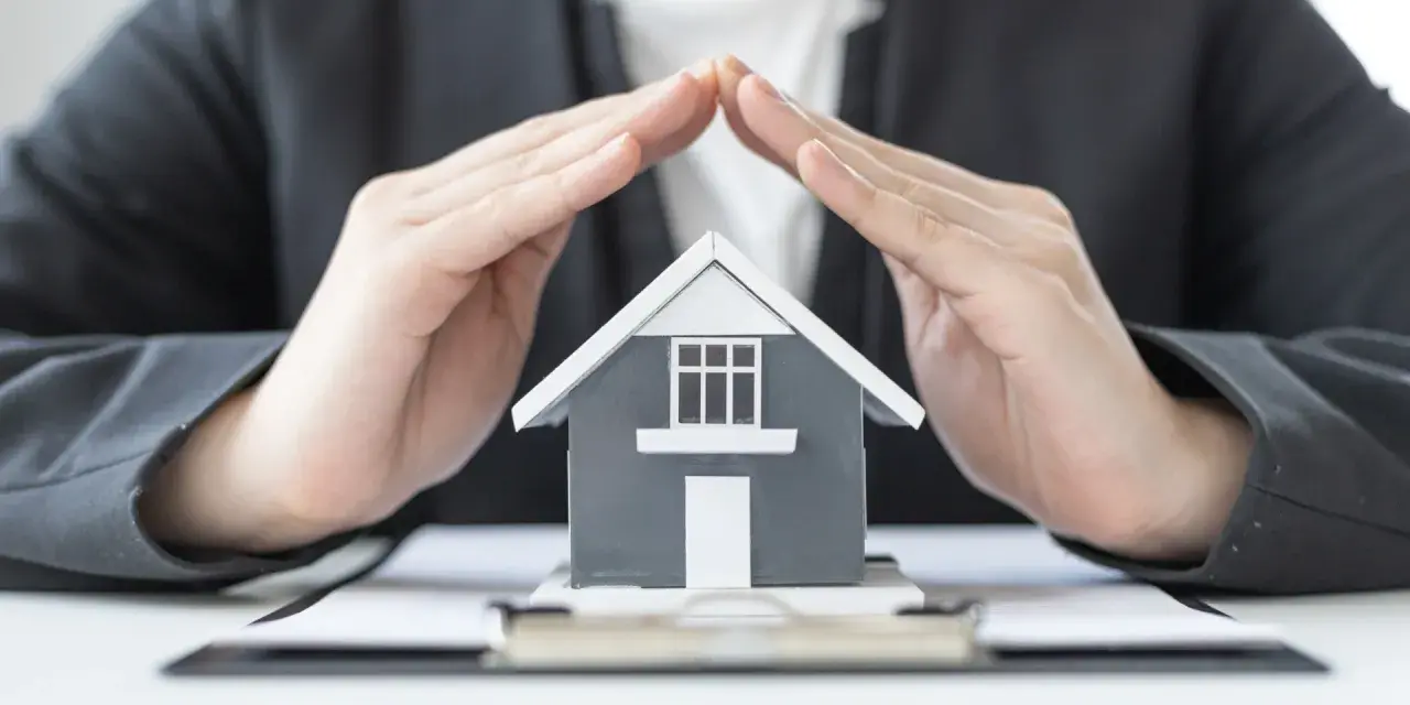 Mains protégeant une maquette de maison, symbolisant la **garantie de bon fonctionnement** et la sécurité de votre investissement immobilier.