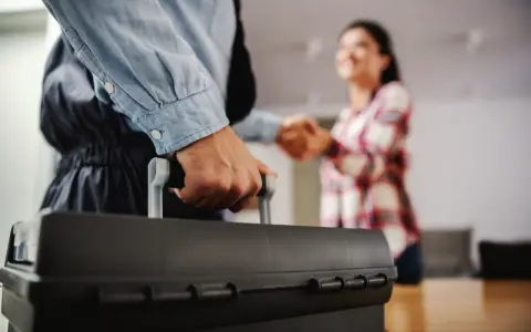 Un plombier arrive avec sa boîte à outils pour régler une fuite d'eau avant compteur sur terrain privé. Une cliente le remercie.