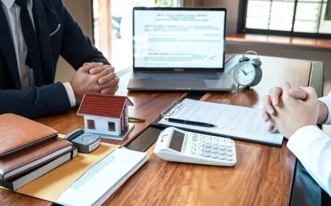 Réunion pour discuter de la faute du mandataire de gestion locative : maison modèle, calculatrice, documents et ordinateur portable sur un bureau en bois.