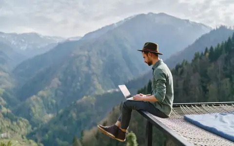 Un homme en chapeau travaille sur son ordinateur portable, profitant du télétravail à l'étranger avec une vue imprenable sur les montagnes.