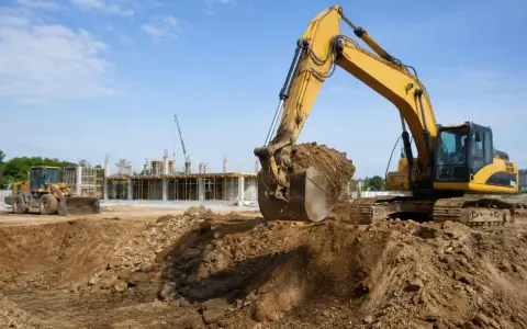 Pelleteuse jaune en action, modifiant le niveau du terrain naturel sur un chantier. Une chargeuse et une grue sont visibles en arrière-plan.