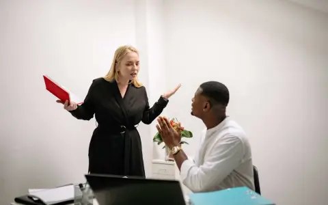 Une femme en colère, tenant un livre rouge, semble se faire insulter au travail par un collègue qui a les mains jointes.