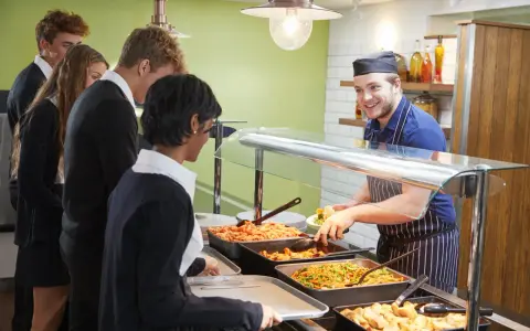Des élèves font la queue pour le déjeuner à la cantine. Un employé souriant sert des légumes. Un contrat extra restauration est en cours de discussion.