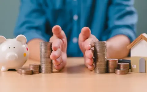 Mains protégeant des piles de pièces, symbolisant la gestion financière et la **desolidarisation pret immo**. Une tirelire et une maison miniature sont visibles.