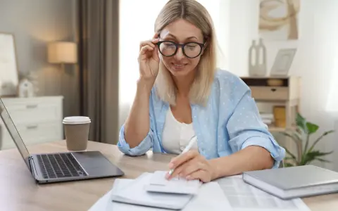 Femme aux lunettes calculant, laptop et café à proximité. Elle semble gérer des finances, peut-être une réduction de capital non motivée par des pertes.