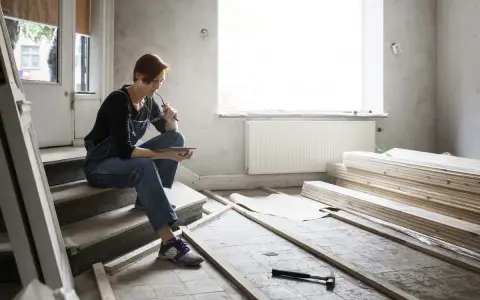 Femme en salopette assise sur des marches, tablette à la main, réfléchissant aux travaux de second œuvre. Matériaux de construction et outils au sol.