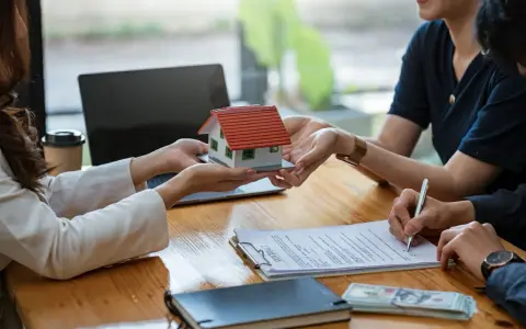 Une maison miniature est remise, un contrat est signé, et de l'argent est posé sur la table, suggérant une transaction immobilière et un chèque de caution.