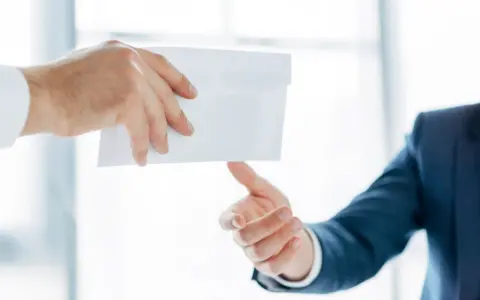Main d'un homme en costume remettant une enveloppe blanche à une autre main. Peut-être une mise en demeure URSSAF avec un délai de paiement.