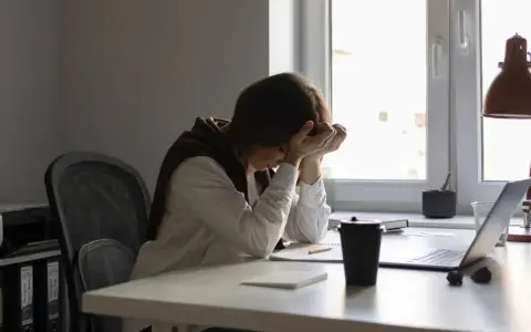 Une personne accablée par la pression au travail, la tête entre les mains, assise à un bureau encombré.