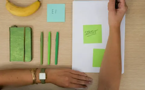 Une main écrit "STATUT" sur un post-it vert, à côté d'un carnet, de stylos et d'une banane. L'acronyme "EI" sur un post-it bleu suggère une **entreprise individuelle**.