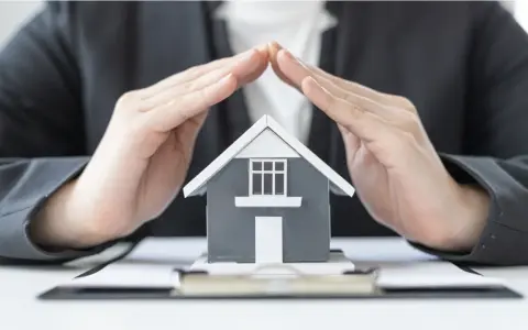Mains protégeant une maquette de maison, symbolisant la **garantie de bon fonctionnement** et la sécurité de votre investissement immobilier.