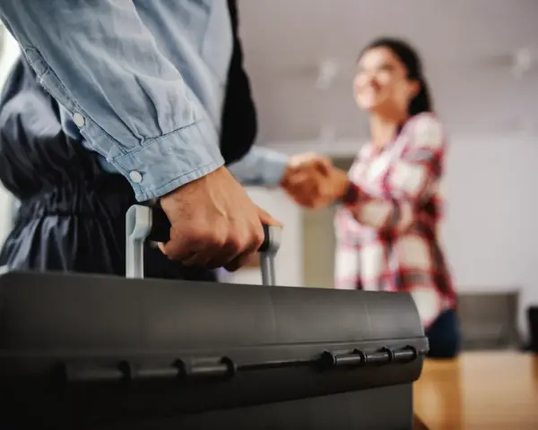 Un plombier arrive avec sa boîte à outils pour régler une fuite d'eau avant compteur sur terrain privé. Une cliente le remercie.