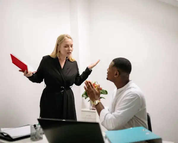Une femme en colère, tenant un livre rouge, semble se faire insulter au travail par un collègue qui a les mains jointes.