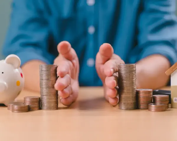 Mains protégeant des piles de pièces, symbolisant la gestion financière et la **desolidarisation pret immo**. Une tirelire et une maison miniature sont visibles.