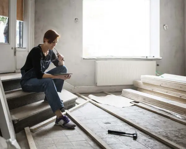 Femme en salopette assise sur des marches, tablette à la main, réfléchissant aux travaux de second œuvre. Matériaux de construction et outils au sol.