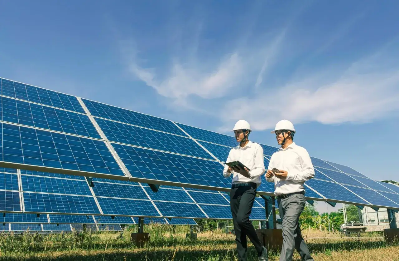 Deux hommes en casque blanc inspectent un champ de panneaux solaires, discutant d'un bail emphytéotique photovoltaïque sous un ciel bleu.