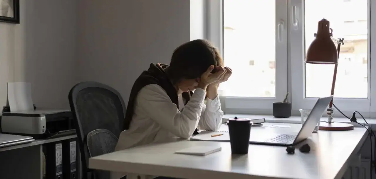 Une employée de bureau, accablée, semble souffrir du burn out fonction publique. Elle est assise à son bureau, la tête entre les mains, devant un ordinateur portable et une tasse.