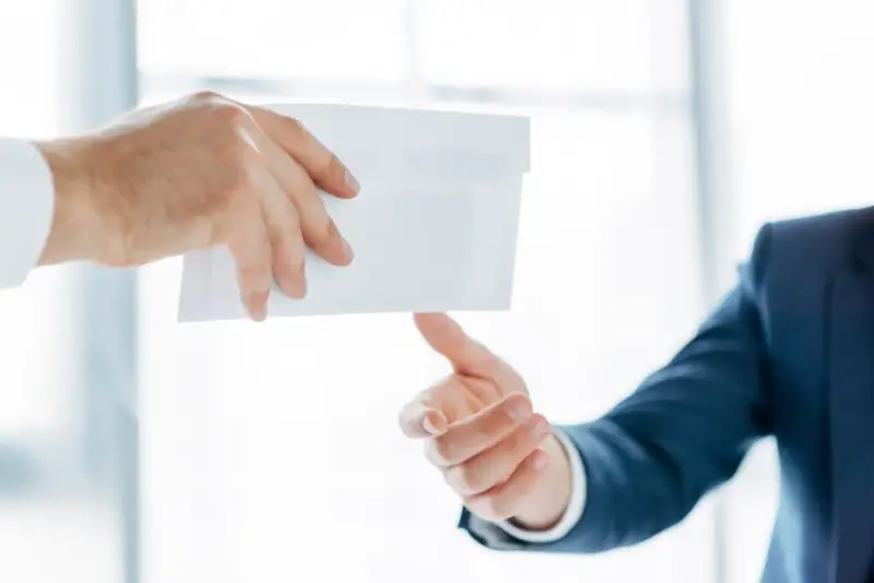 Main d'un homme en costume remettant une enveloppe blanche à une autre main. Peut-être une mise en demeure URSSAF avec un délai de paiement.